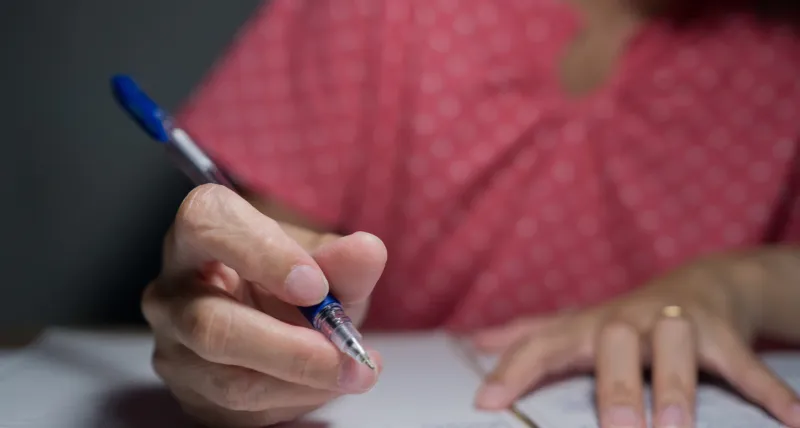 Pessoa escrevendo em um papel com uma caneta azul, representando a participação em provas ou concursos públicos.